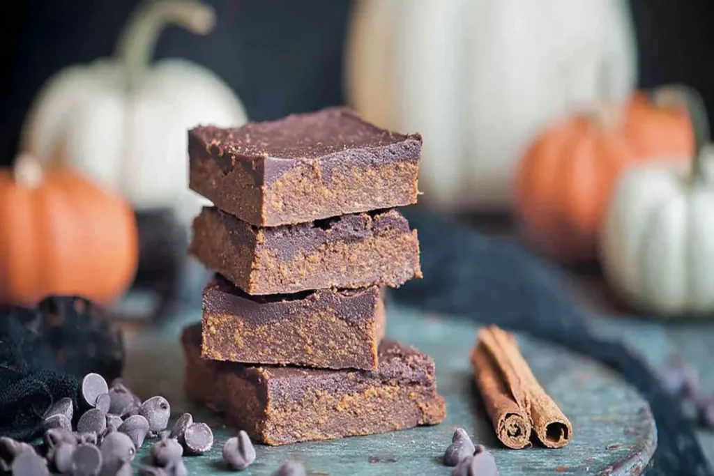 A stack of four rich and moist Healthy Pumpkin Fudge Brownies on parchment paper, surrounded by chocolate chips, cinnamon sticks, and blurred autumn pumpkins.