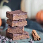 A stack of four rich and moist Healthy Pumpkin Fudge Brownies on parchment paper, surrounded by chocolate chips, cinnamon sticks, and blurred autumn pumpkins.