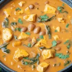 Overhead view of a vibrant bowl of chicken pumpkin soup garnished with fresh herbs, accompanied by lime, ginger, and tortilla chips on a dark background.