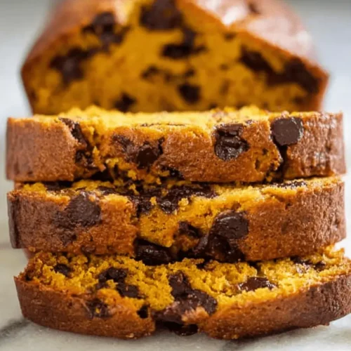 A stack of three moist slices of golden chocolate chip pumpkin bread on a marble surface, with a blurred loaf in the background.