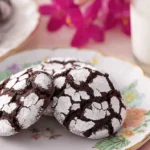 Three chocolate crinkle cookies dusted with powdered sugar on a floral plate, with milk in the background, exuding a warm and cozy aesthetic.