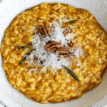 Overhead view of a rustic bowl of Creamy Pumpkin Risotto topped with grated cheese, pecans, and fresh sage.