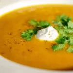 Close-up of a creamy, vibrant curried pumpkin soup recipe garnished with a dollop of cream and fresh herbs in a white patterned bowl.