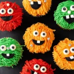 Colorful halloween monster cupcakes with shaggy red, orange, and green frosting, candy eyes, and playful teeth, arranged on a dark surface.