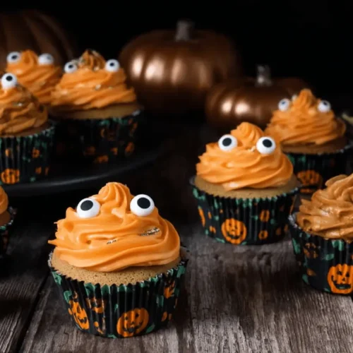 A festive display of orange-frosted halloween pumpkin cupcakes with googly eyes and jack-o'-lantern liners on a rustic wooden table with decorative metallic pumpkins.