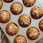 Top-down view of freshly baked healthy pumpkin muffins with oat toppings in a metal pan on a light countertop.