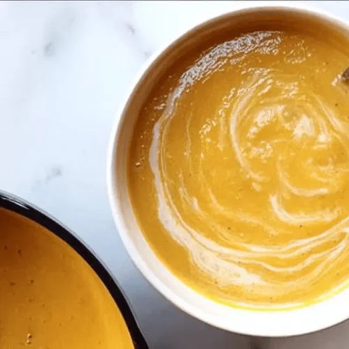 Overhead view of two creamy bowls of Ina Garten pumpkin soup on a white marble surface, garnished with cream swirls and a spoon.