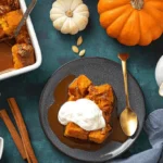 Overhead flat lay of a festive fall dessert: pumpkin bread pudding with caramel sauce and whipped cream, surrounded by small pumpkins and autumn spices.