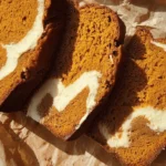 Three moist slices of homemade pumpkin bread with cream cheese swirls, artfully arranged on crumpled parchment paper.