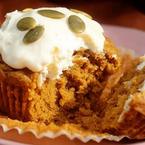 A single, moist pumpkin cream cheese muffins, broken open to show its tender interior, topped with creamy white frosting and green pumpkin seeds on a pale pink plate.