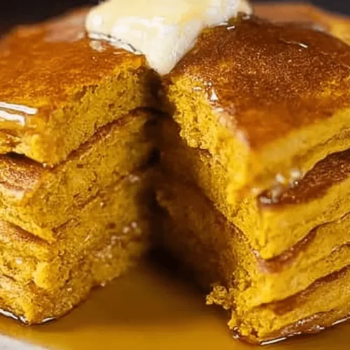 Close-up of a stack of golden-orange pumpkin pancakes topped with melting butter and dripping syrup on a light plate.