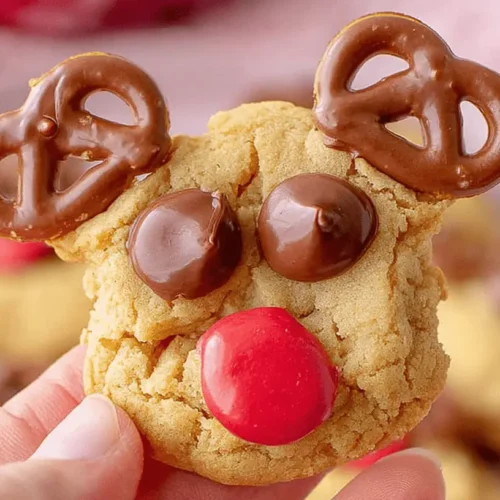 A charming, handmade reindeer cookies with pretzel antlers and a red candy nose, held gently in a hand against a blurred pink background.