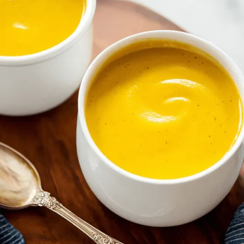 Two mugs of creamy roasted butternut squash soup served on a dark wooden board with an antique spoon.