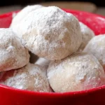 Freshly baked Snowball Cookies, generously coated in powdered sugar, served in a vibrant red bowl on a rustic wooden table.