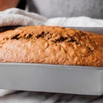 Hands carefully holding a freshly baked, golden-brown loaf of sourdough pumpkin bread in a metal pan on a marble countertop.