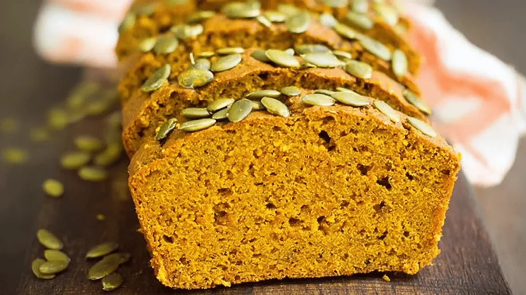Deliciously sliced vegan pumpkin bread loaf, generously topped with vibrant green pumpkin seeds on a rustic wooden board.