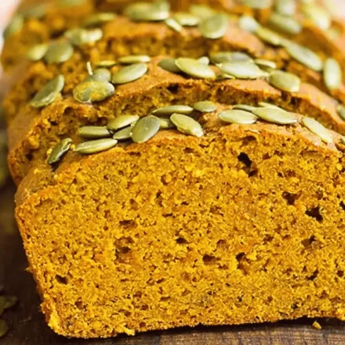Deliciously sliced vegan pumpkin bread loaf, generously topped with vibrant green pumpkin seeds on a rustic wooden board.