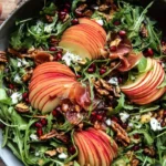 A vibrant Autumn Salad, artfully arranged with arugula, red apple slices, pomegranate arils, pecans, goat cheese, and crispy prosciutto in a rustic bowl on a wooden table.