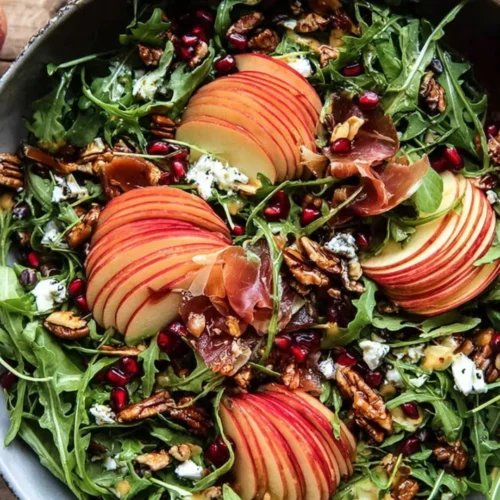 A vibrant Autumn Salad, artfully arranged with arugula, red apple slices, pomegranate arils, pecans, goat cheese, and crispy prosciutto in a rustic bowl on a wooden table.