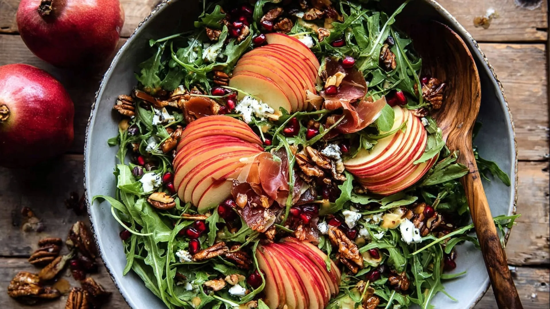 A vibrant Autumn Salad, artfully arranged with arugula, red apple slices, pomegranate arils, pecans, goat cheese, and crispy prosciutto in a rustic bowl on a wooden table.