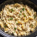 Close-up, top-down view of creamy Crockpot Chicken Pasta with fusilli, shredded chicken, and a rich sauce, garnished with fresh parsley and red pepper flakes in a black slow cooker.
