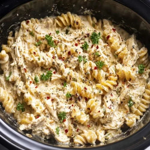 Close-up, top-down view of creamy Crockpot Chicken Pasta with fusilli, shredded chicken, and a rich sauce, garnished with fresh parsley and red pepper flakes in a black slow cooker.