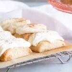 Close-up of homemade Nutmeg Logs cookies on a metal cooling rack lined with parchment paper, showing golden-brown rectangular bars topped with thick creamy white frosting and sprinkled with nutmeg, with a rose-pink glass cake stand and soft white fabric in the background.