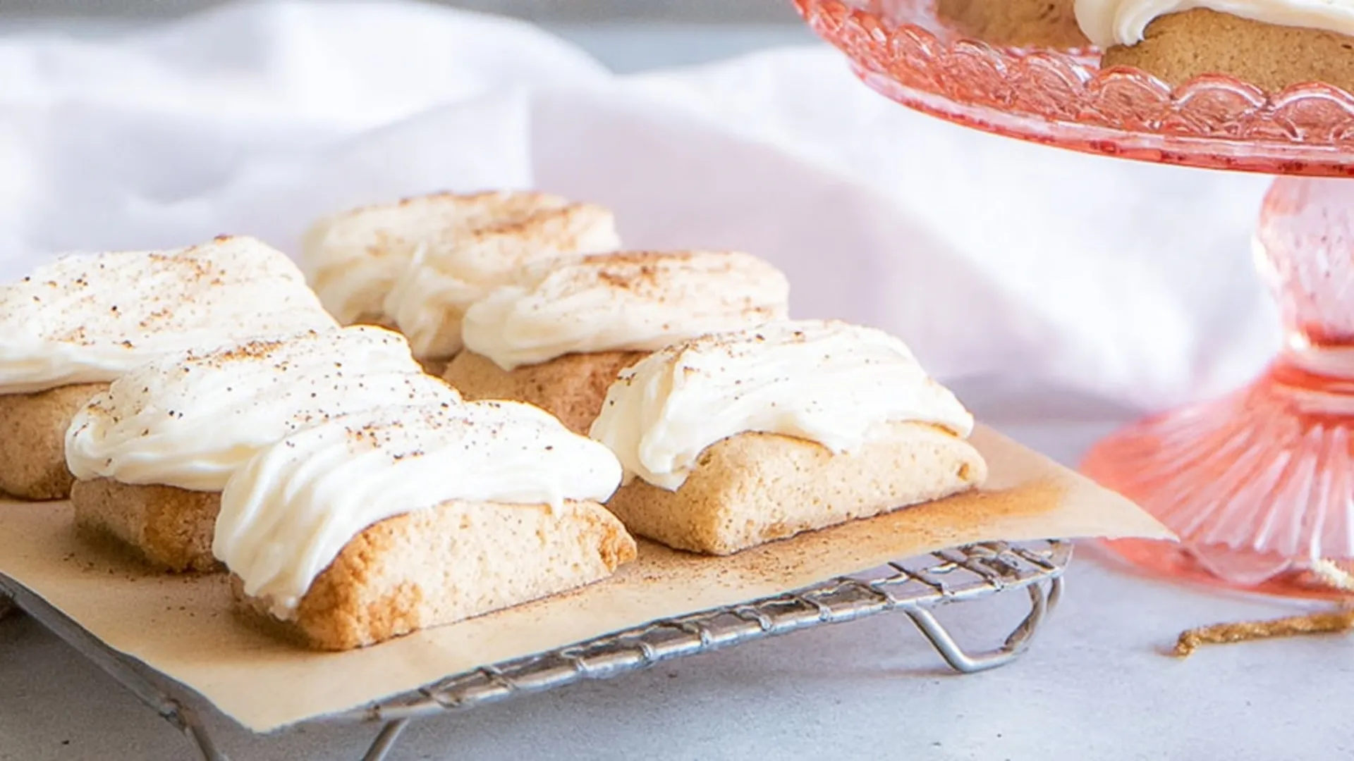 Close-up of homemade Nutmeg Logs cookies on a metal cooling rack lined with parchment paper, showing golden-brown rectangular bars topped with thick creamy white frosting and sprinkled with nutmeg, with a rose-pink glass cake stand and soft white fabric in the background.