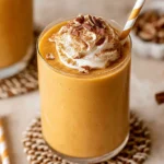 A creamy pumpkin pie smoothie in a clear glass, topped with pecans and spices, set on a light surface with cinnamon sticks.