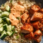 Close-up of a vibrant poke bowl featuring perfectly cooked, cubed Air Fryer Salmon, fresh cucumber, brown rice, and a creamy orange sauce in a blue and white patterned bowl.