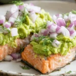 Baked Salmon with Avocado Mash Close-up of two appetizing baked salmon fillets generously topped with vibrant green avocado mash, diced red onion, and fresh dill on a rustic plate.