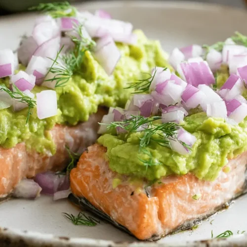 Baked Salmon with Avocado Mash Close-up of two appetizing baked salmon fillets generously topped with vibrant green avocado mash, diced red onion, and fresh dill on a rustic plate.