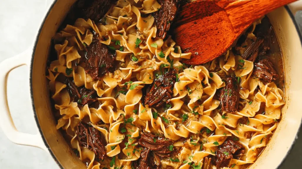 Overhead view of a hearty beef and noodles dish with shredded beef and golden egg noodles in a cream-colored pot, garnished with fresh parsley and a wooden spoon.