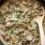 Top-down view of a hearty beef stroganoff in a rustic blue-handled pot, garnished with fresh chives.