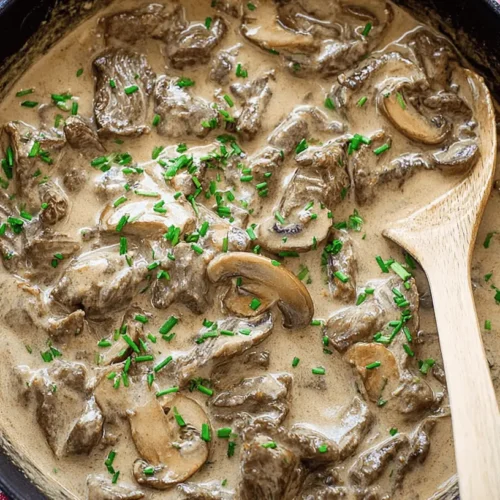 Top-down view of a hearty beef stroganoff in a rustic blue-handled pot, garnished with fresh chives.