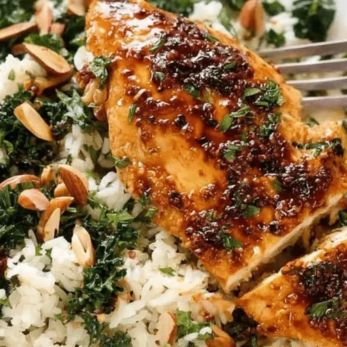 Close-up of glazed chicken breast served with white rice, green vegetables, and toasted almonds on a white plate.