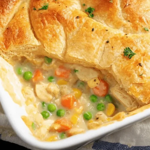 Freshly baked chicken pot pie casserole in a white dish, featuring a golden, flaky puff pastry crust and a visible creamy filling with chicken, peas, and carrots.