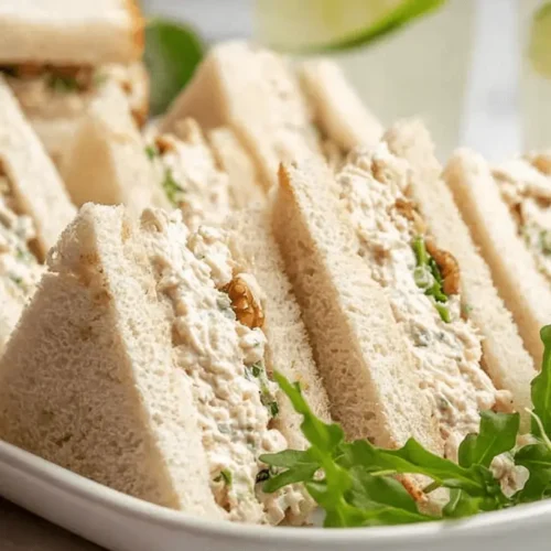 Platter of elegant chicken sandwiches with creamy herb and nut filling, garnished with arugula.