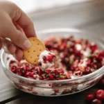 A hand dips a cracker into a festive Christmas Cranberry Jalapeno Dip, garnished with fresh cranberries and herbs, on a wooden table.