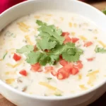 A vibrant close-up of creamy Christmas Queso dip in a white bowl, garnished with cilantro and diced tomatoes, surrounded by golden tortilla chips on a rustic wooden surface.