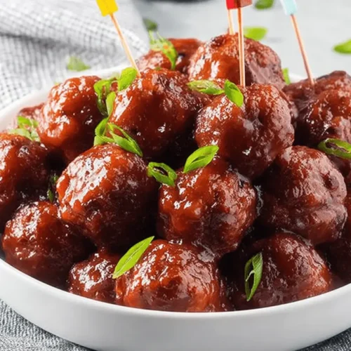 Close-up of glistening Cranberry Meatballs in a white bowl, garnished with fresh green herbs and colorful toothpicks, ready to serve.