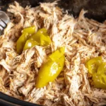 Close-up of tender, shredded Crockpot Mississippi Chicken with bright yellow-green peppers and serving utensils in a dark pot.