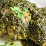 Close-up of succulent Crockpot Pesto Chicken pieces, richly coated in vibrant green pesto sauce, garnished with fresh herbs on a white plate.