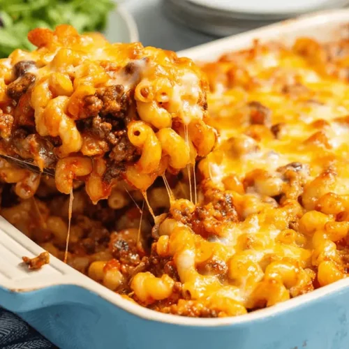 Close-up of a serving spoon lifting a cheesy portion of a baked macaroni and ground beef casserole recipe from a blue ceramic dish.