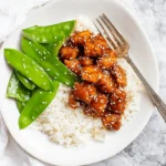 Overhead view of a delicious plate of healthy sesame chicken, fluffy white rice, and bright green snow peas, garnished with sesame seeds and an elegant fork.