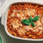 A top-down view of a freshly baked, bubbling High Protein Spaghetti Casserole with melted cheese, fresh basil, and chili flakes in a white ceramic dish on a speckled countertop.
