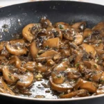 Close-up overhead shot of glistening Marsala Mushrooms simmering in a dark non-stick pan, with garlic and fresh herbs.