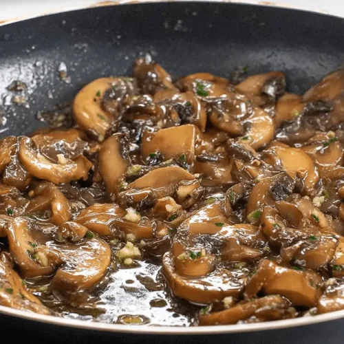 Close-up overhead shot of glistening Marsala Mushrooms simmering in a dark non-stick pan, with garlic and fresh herbs.