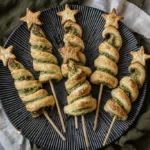 Six golden-brown Puff Pastry Pesto Trees, shaped like Christmas trees with star toppings, arranged on a textured dark plate with festive lights.