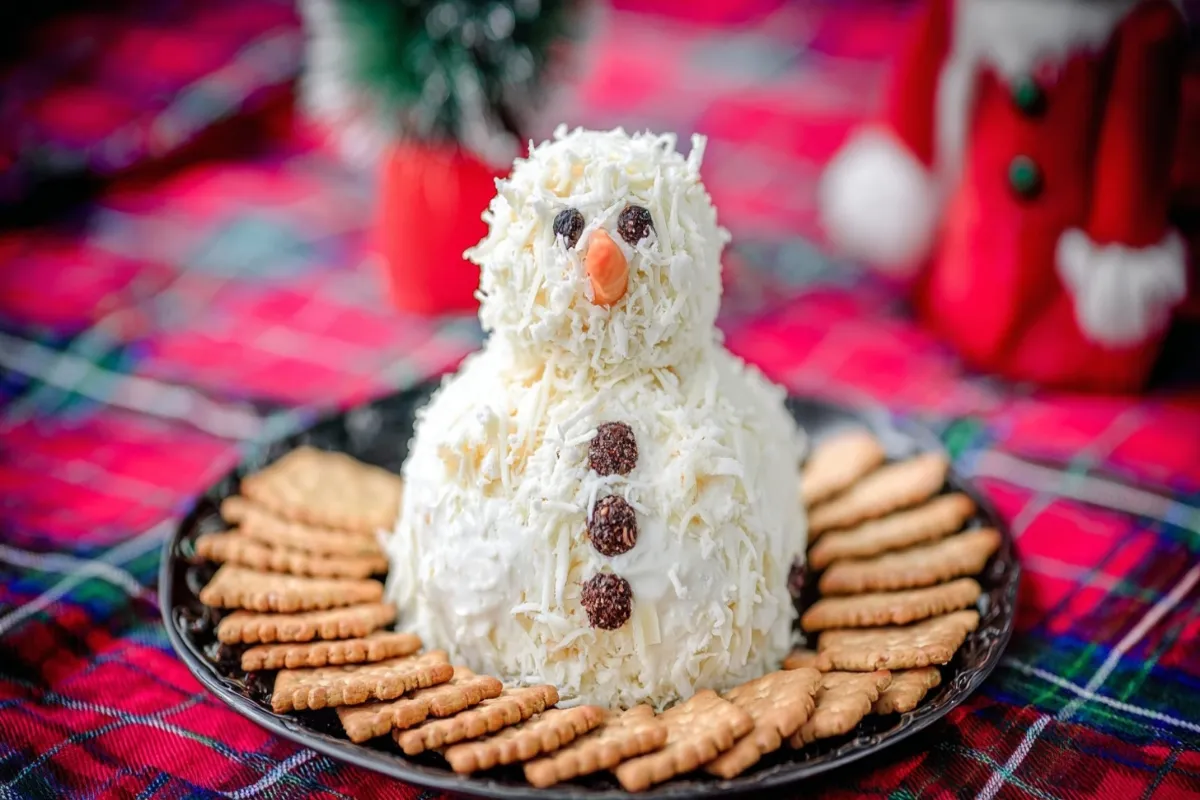 Festive Ranch Snowman Cheeseball, adorned with a top hat and facial features, served with crackers on a holiday plaid background.